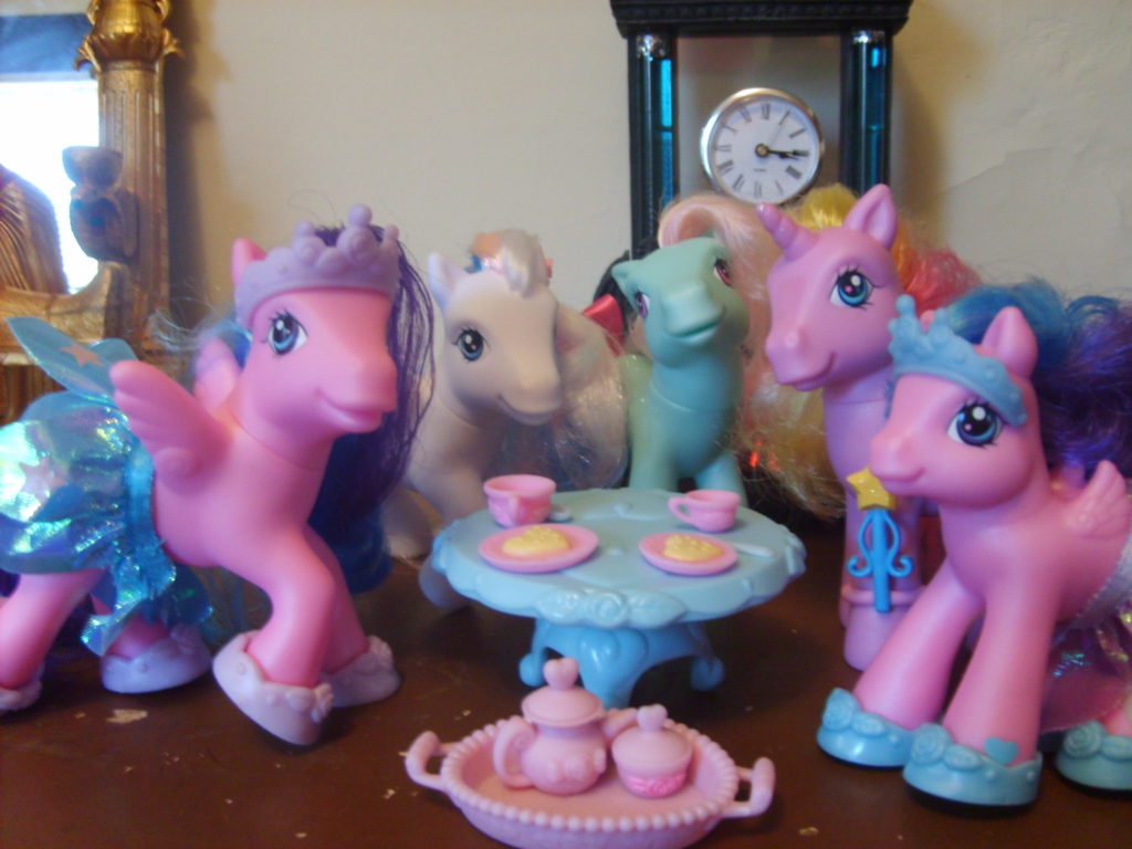 Five little ponies in different colors standing around a table with pastries and cups of tea